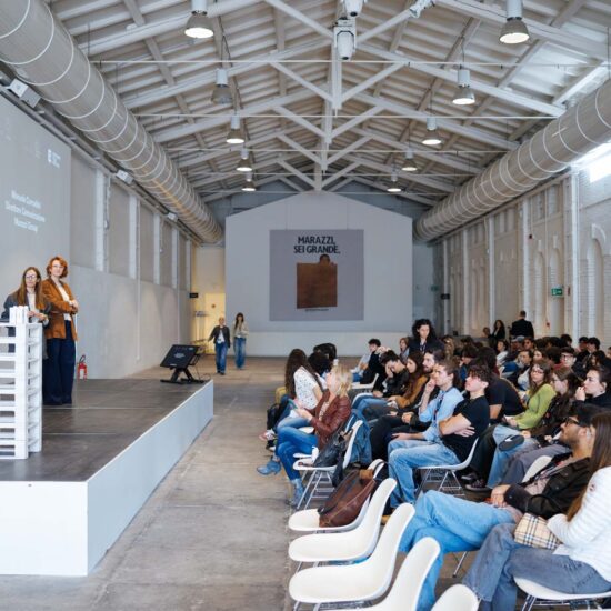 Ambiente interno di un auditorium con pubblico in attesa, palco in legno chiaro e pareti bianche, atmosfera professionale.